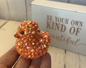 May include: A small, orange rubber ducky covered in iridescent rhinestones. The ducky has a black eye and a small, orange beak.