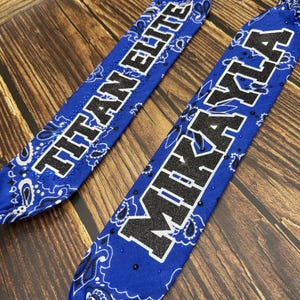 Könnte beinhalten: Zwei blaue Bandanas mit weißen Paisley-Mustern und den Texten "TITAN ELITE" und "MIKAYLA" in Schwarz und Weiß. Die Bandanas sind an den Enden geknotet und liegen auf einer Holzoberfläche.