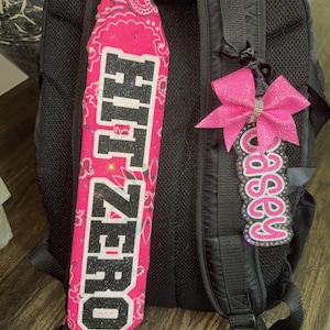 Puede incluir: Mochila negra con dos accesorios decorativos. Uno es un pañuelo rosa con el texto "HIT ZERO" en purpurina negra. El otro es un lazo rosa con una etiqueta con el nombre "Casey" en rosa y blanco.