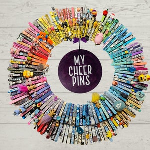 Può includere: Una ghirlanda colorata realizzata con mollette per bucato con parole e frasi scritte su di esse. La ghirlanda è decorata con piccoli giocattoli e un cerchio viola al centro che dice "I miei Cheer Pins".