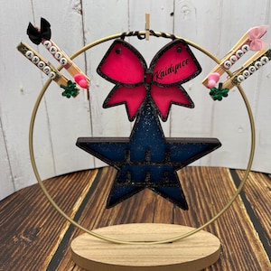 Könnte beinhalten: Ein goldfarbener Metallständer mit runder Oberseite und drei Beinen. Ein blauer Stern mit Glitzer und eine rosafarbene Schleife mit Glitzer hängen am Ständer. Die Schleife trägt den Namen "Kaidynce". Zwei Holz-Wäscheklammern mit den Worten "Slay" und "Lucky" sind am Ständer befestigt.