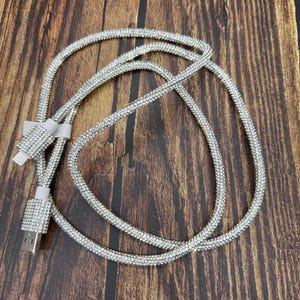 Pode incluir: Um cabo de carregamento prateado com um design de strass transparente. O cabo está enrolado e as pontas são visíveis.