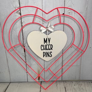 May include: A pink metal heart-shaped wreath with a white heart-shaped sign that says "My Cheer Pins" hanging from it.