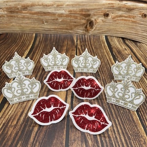 May include: A collection of decorative embellishments, featuring white and gold glitter crowns and red glitter lips. The items are arranged on a wooden surface. These items are suitable for crafting or party decorations. The crowns and lips are eye-catching.