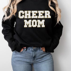 May include: Black sweatshirt with the text "CHEER MOM" in white letters with a gold outline. The sweatshirt is paired with light blue jeans. The overall style is casual and comfortable.