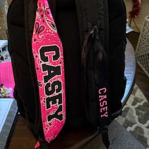 Op de afbeelding: Een zwarte rugzak met twee gepersonaliseerde accessoires. Eén is een felroze bandana met de naam "CASEY" in zwarte blokletters, en de andere is een zwarte band met de naam "CASEY" in roze letters.