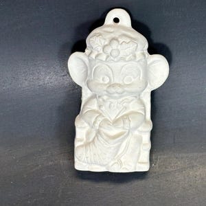 Puede incluir: Un adorno de cerámica blanca que representa un ratón de dibujos animados con un sombrero adornado con acebo y sentado en una silla. El adorno tiene orejas grandes y un agujero en la parte superior para colgar. El ratón lleva un vestido.