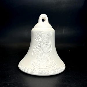 May include: A white ceramic bell featuring an embossed angel design holding a candle. The bell has a flared base and a loop at the top for hanging. The angel design includes wings and a patterned dress.