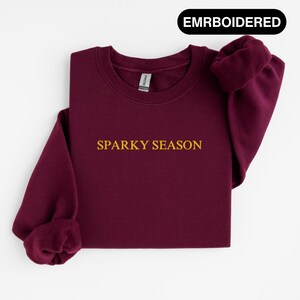 Könnte beinhalten: Ein weinrotes Sweatshirt mit dem gestickten Text "SPARKY SEASON" in goldfarbenen Buchstaben. Das Sweatshirt hat einen Rundhalsausschnitt und lange Ärmel mit gerippten Bündchen. Das Wort "EMRBOIDERED" ist in einem schwarzen Rechteck oben aufgedruckt.