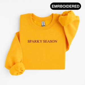Könnte beinhalten: Senfgelbes Sweatshirt mit dem Text "SPARKY SEASON" in dunkelroter Schrift. Das Sweatshirt hat einen Rundhalsausschnitt und lange Ärmel mit umgeschlagenen Bündchen. Das Wort "EMRBOIDERED" befindet sich in einem schwarzen Oval.