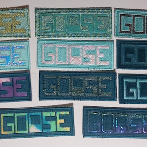 May include: A collection of embroidered patches with the word "GOOSE" in various colors. The patches feature a rectangular shape with the word in a block letter style. Colors include teal, blue, and iridescent hues.
