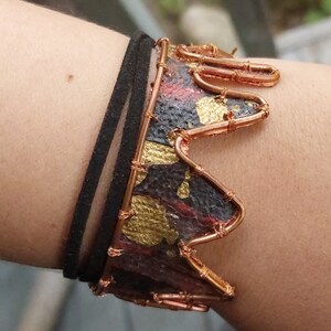 May include: A copper wire bracelet with a crown design. The crown is made of copper wire and has a black and gold patterned fabric inside. The bracelet is worn on a person's wrist.