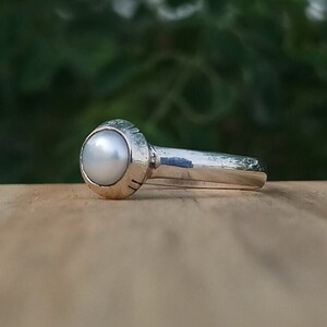 Könnte beinhalten: Ein silberner Ring mit einem weißen Perlmutt, das in eine runde Fassung gefasst ist. Der Ring hat ein strukturiertes Band.
