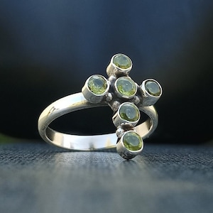 Könnte beinhalten: Ein silberner Ring mit einem Kreuzdesign, das mit grünen Edelsteinen besetzt ist. Das Kreuz besteht aus sieben runden Steinen, die in eine silberne Band gefasst sind.