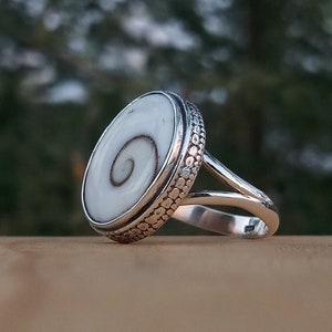 May include: A silver ring with a white shell inlay featuring a spiral pattern. The ring band is textured with small dots.