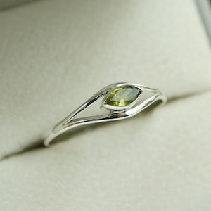 Puede incluir: Un anillo de plata con una gema verde oliva en forma de marquesa. La banda del anillo tiene un diseño fluido y abierto que acuna la piedra. El anillo se presenta sobre un fondo neutro, resaltando su elegante simplicidad.