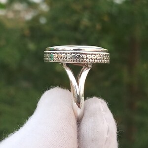Op de afbeelding: Een zilveren ring met een ronde, getextureerde bovenkant en een decoratieve band. De ring ligt op een wit oppervlak.