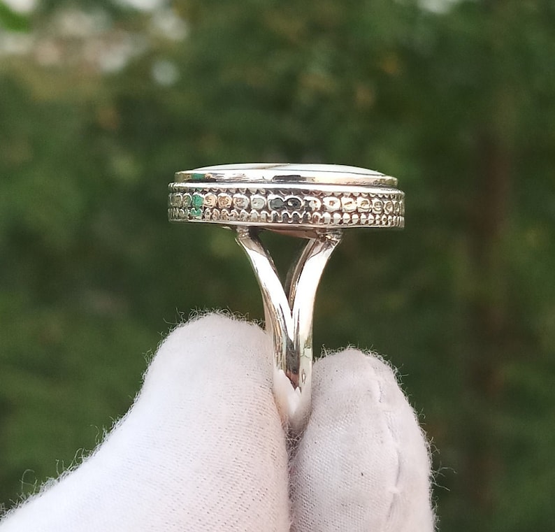 Op de afbeelding: Een zilveren ring met een ronde, getextureerde bovenkant en een decoratieve band. De ring ligt op een wit oppervlak.