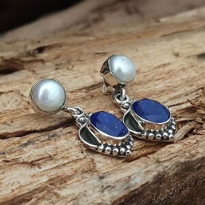 Pode incluir: Um par de brincos de prata com uma pedra de lapis-laz&uacute;li azul e uma p&eacute;rola branca. Os brincos t&ecirc;m o formato de um cora&ccedil;&atilde;o.