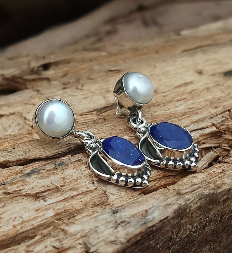 Pode incluir: Um par de brincos de prata com uma pedra de lapis-laz&uacute;li azul e uma p&eacute;rola branca. Os brincos t&ecirc;m o formato de um cora&ccedil;&atilde;o.