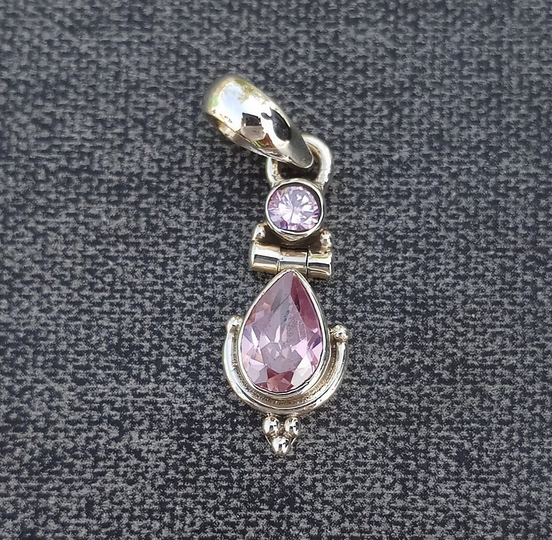 May include: A silver pendant with a teardrop-shaped pink gemstone and a smaller round pink gemstone. The pendant has a decorative design with small beads.