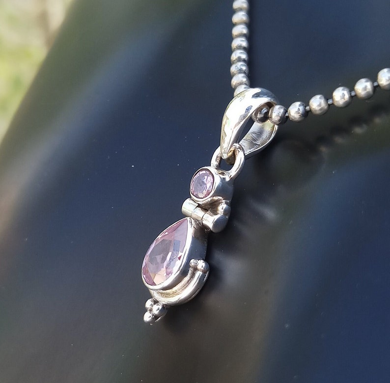 May include: A silver pendant necklace with a teardrop-shaped pink gemstone and a smaller round pink gemstone. The necklace is on a silver chain.