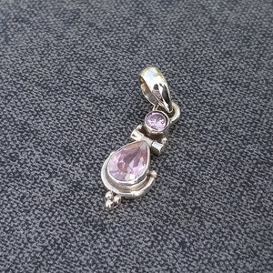 May include: A silver pendant with two pink gemstones. The larger gemstone is a teardrop shape and the smaller gemstone is round. The pendant has a detailed design with small beads and swirls.