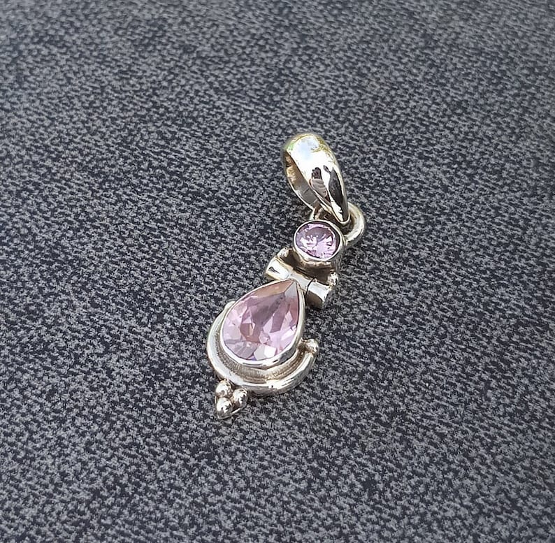 May include: A silver pendant with two pink gemstones. The larger gemstone is a teardrop shape and the smaller gemstone is round. The pendant has a detailed design with small beads and swirls.