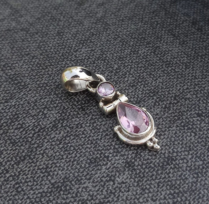 May include: A silver pendant with two pink gemstones. The larger gemstone is a teardrop shape and the smaller gemstone is round. The pendant has a bail for attaching to a chain.