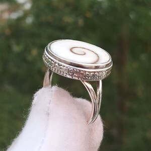 Op de afbeelding: Een zilveren ring met een witte en bruine schelp inlay. De schelp heeft een spiraalvormig patroon. De ring heeft een getextureerde band.