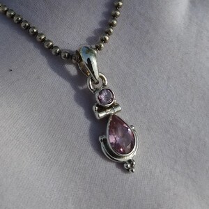 May include: A silver necklace with a pendant featuring two pink gemstones. The larger gemstone is a teardrop shape and the smaller gemstone is round.