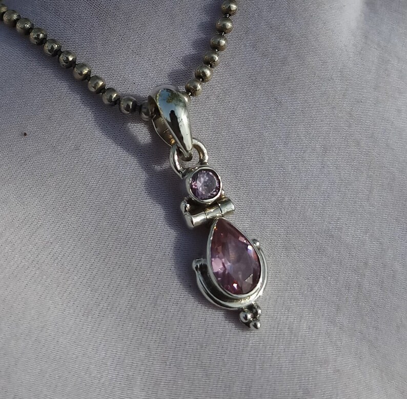 May include: A silver necklace with a pendant featuring two pink gemstones. The larger gemstone is a teardrop shape and the smaller gemstone is round.