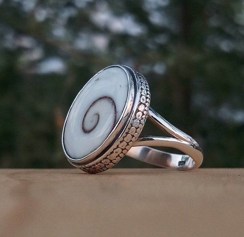 Op de afbeelding: Een zilveren ring met een witte schelp inlay met een spiraalvormig patroon. De ringband is getextureerd met kleine stippen.