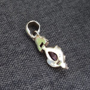 May include: Silver pendant with a purple gemstone set in a detailed design. The pendant has a loop at the top for attaching to a chain.