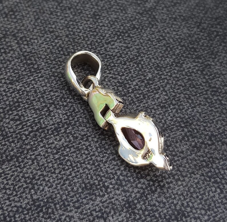 May include: Silver pendant with a purple gemstone set in a detailed design. The pendant has a loop at the top for attaching to a chain.