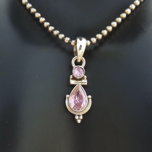 May include: A silver necklace with a pendant featuring a teardrop-shaped pink gemstone and a smaller round pink gemstone. The pendant has a delicate, ornate design.