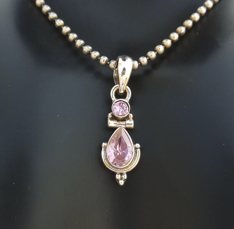 May include: A silver necklace with a pendant featuring a teardrop-shaped pink gemstone and a smaller round pink gemstone. The pendant has a delicate, ornate design.