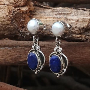 Pode incluir: Um par de brincos de prata com uma p&eacute;rola branca e uma pedra de l&aacute;pis-laz&uacute;li azul. Os brincos t&ecirc;m um design delicado com um pequeno pingente de prata.