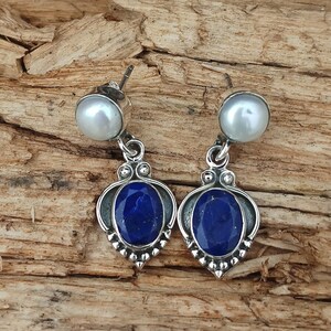Pode incluir: Um par de brincos de prata com uma pedra de l&aacute;pis-laz&uacute;li azul e uma p&eacute;rola branca. Os brincos t&ecirc;m um design delicado e ornamentado.