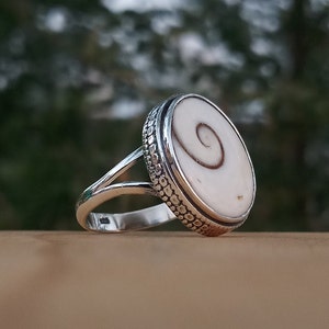Op de afbeelding: Een zilveren ring met een witte schelp inlay met een spiraalvormig patroon. De ring heeft een getextureerde band.