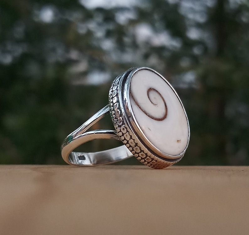 Op de afbeelding: Een zilveren ring met een witte schelp inlay met een spiraalvormig patroon. De ring heeft een getextureerde band.