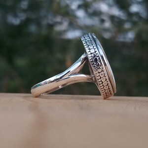 Op de afbeelding: Een zilveren ring met een witte ovale steen gezet in een getextureerde bezel. De ringband is zilver en heeft een delicaat, gedraaid ontwerp.