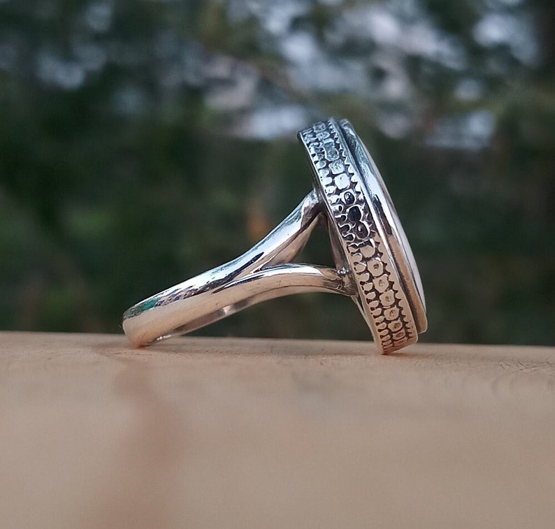 Op de afbeelding: Een zilveren ring met een witte ovale steen gezet in een getextureerde bezel. De ringband is zilver en heeft een delicaat, gedraaid ontwerp.