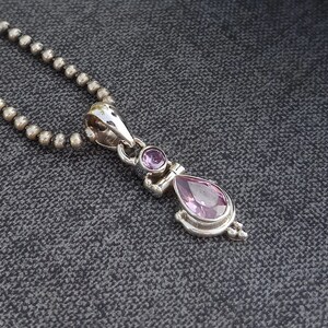 May include: A silver necklace with a pendant featuring two pink gemstones. The pendant is shaped like a teardrop with a smaller round gemstone above it.