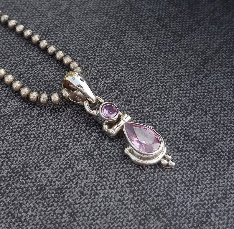 May include: A silver necklace with a pendant featuring two pink gemstones. The pendant is shaped like a teardrop with a smaller round gemstone above it.