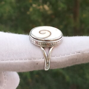 Op de afbeelding: Een zilveren ring met een witte schelp inlay met een spiraalvormig ontwerp. De ring heeft een decoratieve band met een textuurpatroon.