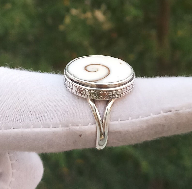 Op de afbeelding: Een zilveren ring met een witte schelp inlay met een spiraalvormig ontwerp. De ring heeft een decoratieve band met een textuurpatroon.