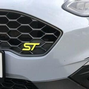 Peut inclure: Gros plan sur la calandre avant d'une voiture grise avec un motif en nid d'abeille noir. La calandre présente un emblème "ST" jaune vif. Le phare de la voiture et une partie du pare-chocs sont visibles.