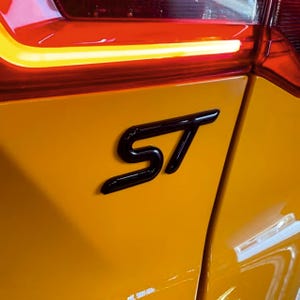May include: Close-up of a yellow car panel with a black "ST" emblem. The emblem is glossy and reflects the surrounding light. The car's taillight is visible in the background, with a red and yellow glow.