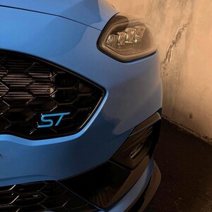 Peut inclure: Gros plan sur l'avant d'une voiture bleu clair. La calandre noire porte les lettres bleues "ST". Le phare est visible à droite. Le pare-chocs de la voiture est noir.